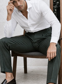 A man wearing a crisp white long-sleeve dress shirt and dark charcoal gray trousers, seated on a wooden chair.