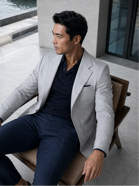 A man wearing a light gray blazer, dark navy polo shirt, and navy trousers sits on a wooden chair in a modern outdoor setting.