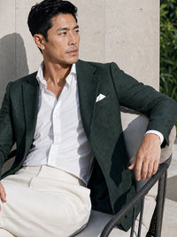 A man wearing a dark green textured blazer over a white button-down shirt and cream trousers, seated on a modern outdoor chair against a light stone wall.