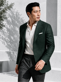 A man wearing a dark forest green blazer over a white polo shirt and dark trousers, standing in a modern outdoor setting with light gray concrete steps and soft shadows.
