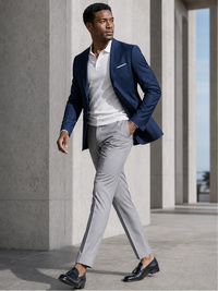 A man walking outdoors in a stylish outfit consisting of a navy blue blazer, white polo shirt, light gray trousers, and black loafers, set against a modern concrete architectural background.