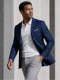 A man wearing a navy blue blazer over a white polo shirt and light gray trousers, standing against a gray concrete wall.