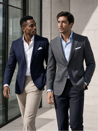 Two men walking outdoors in tailored suits. The man on the left wears a navy blazer with beige trousers and a white shirt, and the man on the right wears a gray blazer with dark navy trousers and a light blue shirt.