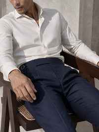 A man seated on a wooden chair wearing a white long-sleeve button-up shirt and dark navy trousers, photographed in warm natural light against a textured wall.