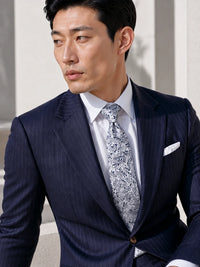 A man wearing a dark navy blue pinstripe suit jacket, a white dress shirt, and a patterned silver and white paisley tie, with a white pocket square.
