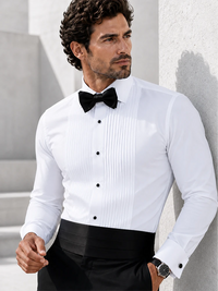 A man wearing a white long-sleeve pleated formal shirt with a black bow tie and black trousers, leaning against a light gray wall.