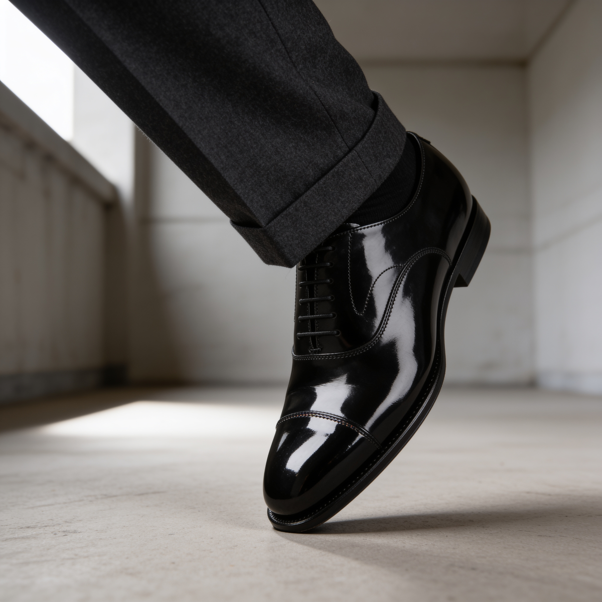 Mirror-polished black cap-toe oxford shoe against the clean break of a dark charcoal wool trouser, low-angle editorial detail shot