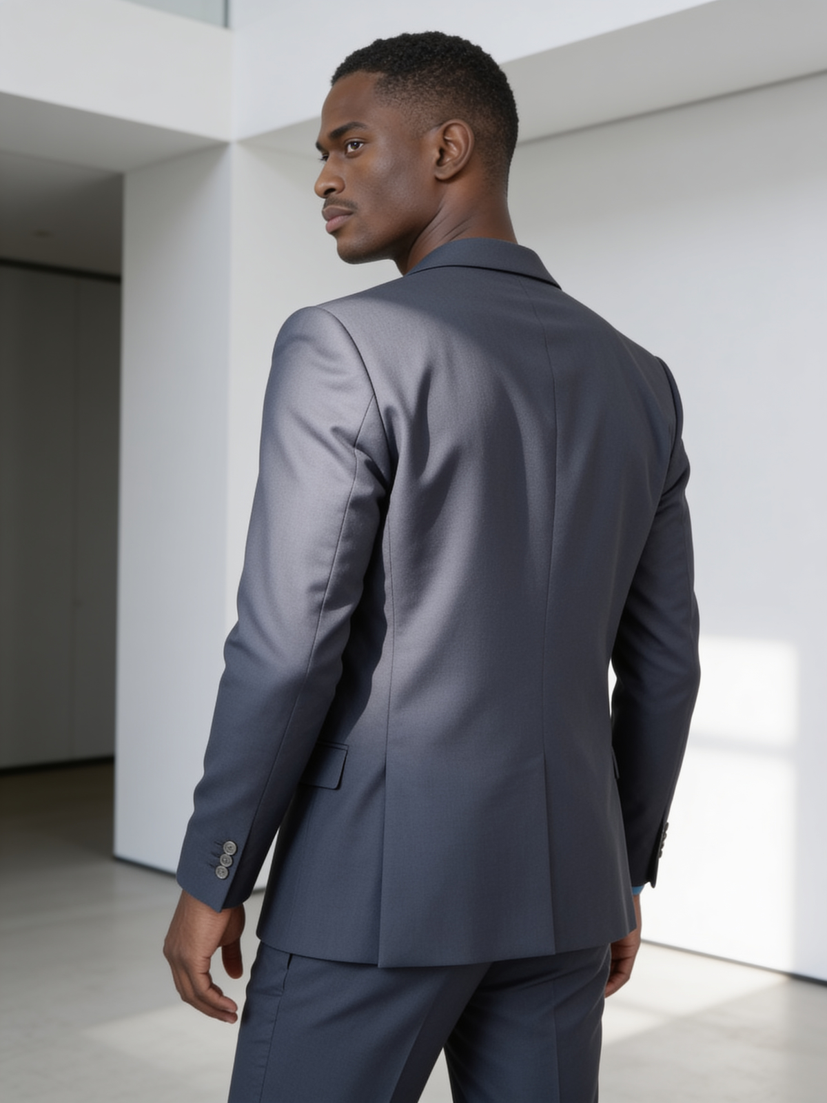Clean single-breasted suit in mid-gray, three-quarter back view showing jacket drape and shoulder line in minimalist architectural interior