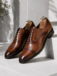 A pair of men's brown leather oxford shoes with cap toes and laces, displayed on wooden shoe trees against a light gray stone background.