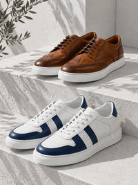 Two pairs of low-top sneakers displayed on light stone steps: brown leather lace-up shoes in the back and white sneakers with navy side stripes in the front. A leafy branch hangs in the upper left, and shadows from leaves are cast across the scene.