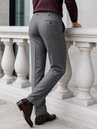 A person wearing gray tailored trousers, a burgundy long-sleeve top, and dark brown dress shoes, standing beside a light stone balustrade.