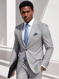 A man wearing a light gray two-piece suit, white dress shirt, and light blue tie, holding a black folder. The background is a clean, modern white architectural setting.