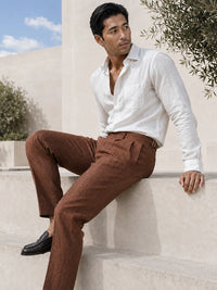 A man wearing a white linen button-down shirt and brown linen trousers sits on outdoor stone steps under a blue sky.