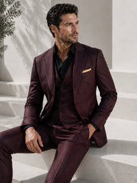 A man wearing a three-piece burgundy suit with a black shirt and a gold pocket square, sitting on light-colored stone steps.