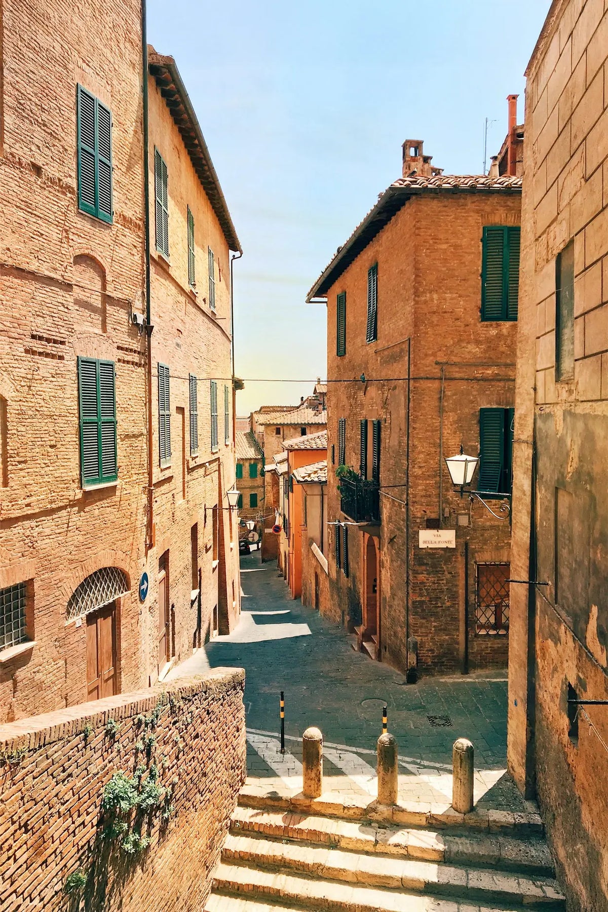 Tight street between brick buildings with green shutters in a sunlit town.