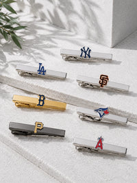 Six silver-toned baseball and sports clip hair ties with team logos, arranged on a light textured surface with soft shadows and a small plant leaf in the corner.
