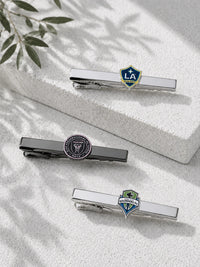 Three silver-toned tie bars with sports team emblems are arranged on a textured white surface with soft leaf shadows. One tie bar features a Los Angeles Galaxy shield, one a circular black-and-gold emblem, and one a Seattle Sounders FC shield.