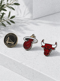 Three sports enamel pins from the Golden State Warriors, Miami Heat, and Chicago Bulls arranged on a light gray textured surface with soft shadows and a plant leaf in the corner.