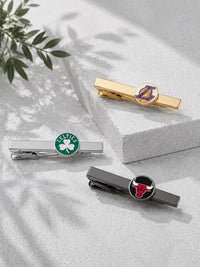 Three tie bars with sports team logos are arranged on a light gray textured surface with soft leaf shadows. One gold tie bar has a Los Angeles Lakers logo, one silver tie bar has a Boston Celtics logo with a green shamrock, and one dark gray tie bar has a Chicago Bulls logo.
