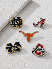 Five collegiate logo enamel pins arranged on a textured white surface: Notre Dame, Alabama, Texas Longhorns, Michigan, and Ohio State. The pins feature gold, red, orange, and navy colors with silver metal outlines.