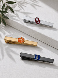 Three metal tie clips arranged on a light textured surface with leaf shadows. The top clip is silver with a red Oklahoma 'OU' logo, the middle clip is gold with an orange paw print, and the bottom clip is black with a blue 'I' logo.