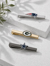 Three silver-tone tie bars with sports team logos, including the New England Patriots, Green Bay Packers, and Dallas Cowboys, arranged on a textured white block with soft leaf shadows.