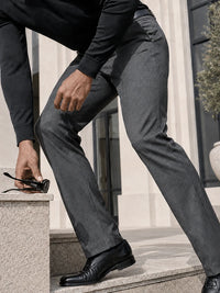 A man wearing slim-fit charcoal gray dress pants, a black long-sleeve shirt, and black leather dress shoes, leaning over stone steps while holding a pair of sunglasses.
