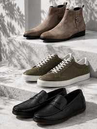 Three pairs of men's casual footwear arranged on light stone steps: taupe suede ankle boots, olive green suede sneakers with white laces and soles, and black leather penny loafers.