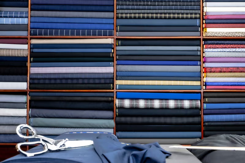 A display of folded fabric bolts arranged in wooden racks, showcasing a wide variety of colors and patterns including plaid, stripes, and solid blues, grays, and whites. A pair of silver tailor's shears and a folded blue fabric piece rest on a table in the foreground.
