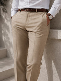 A person wearing beige chinos with a brown leather belt, styled with a white dress shirt and a brown leather watch, leaning against a light stone wall.