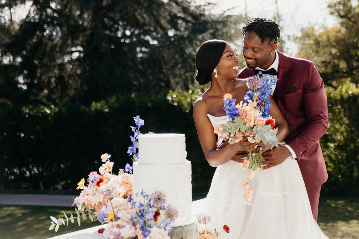 wedding couple embracing next to their wedding cake. groom in burgundy tuxedo