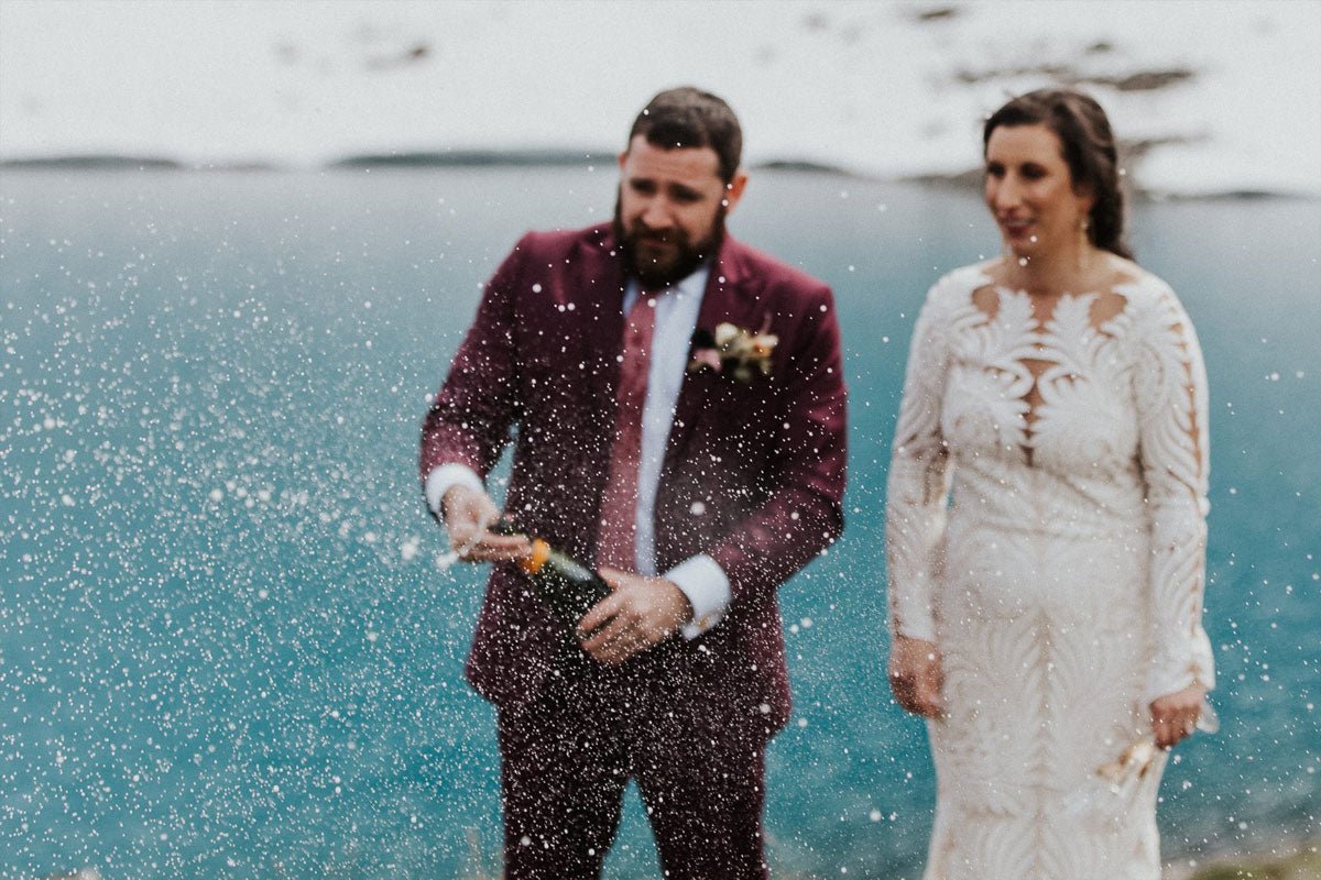 groom popping open a bottle of champagne while his bride looks on