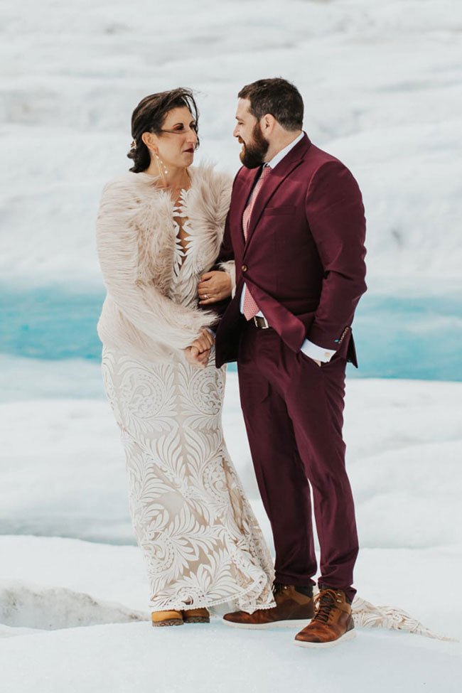 bride and groom in burgundy suit standing on a glacier